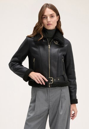 Giacca di pelle nera con colletto e polsini in shearling, zip dorate, vita aderente e dettaglio della cintura. Abbinata a pantaloni grigi a vita alta.