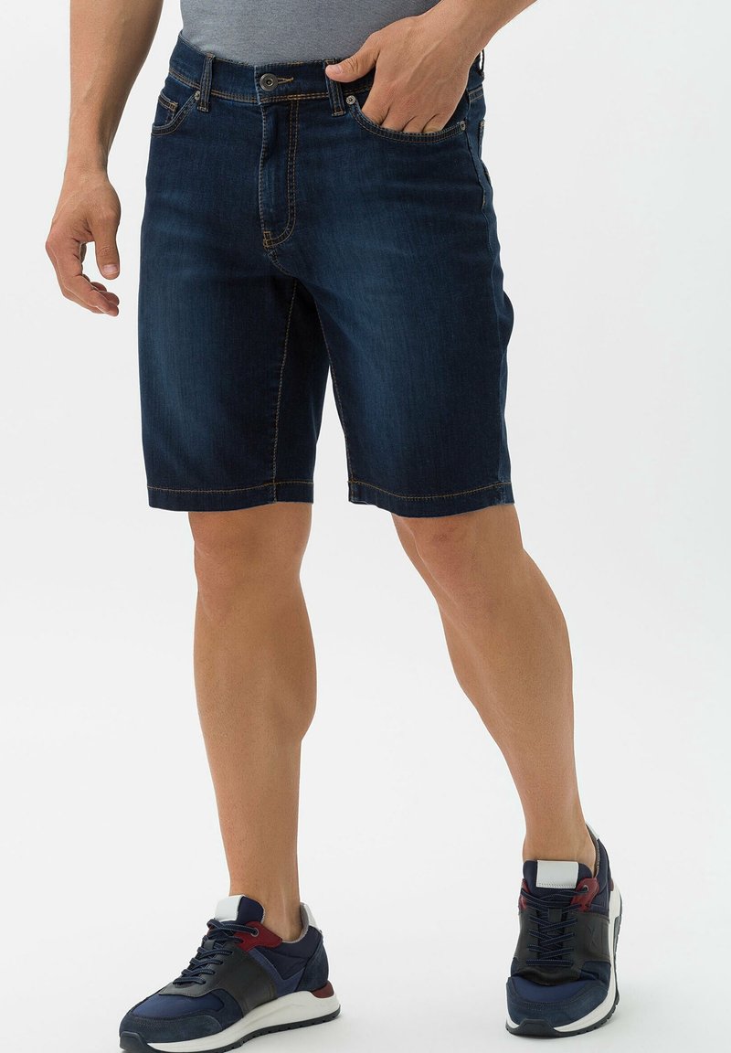 Tummansinisiä farkkuhousuja keltaisilla ompeleilla, joissa on etutaskut, nappikiinnitys ja suora lahkeensuun malli. Yhdistettynä laivastonsinisiin lenkkareihin.