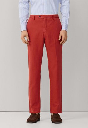 Hombre con pantalones rectos de color óxido, camisa de rayas azul claro metida por dentro y mocasines marrones de ante, de pie frente a un fondo sencillo.