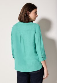 Blusa di lino turchese con maniche a tre quarti, dettaglio con piega sul retro e bottoni decorativi sulle maniche, abbinata a jeans scuri.