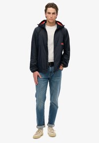 Chaqueta rompevientos en azul marino con un interior rojo, cremallera frontal y mangas largas. Combinada con jeans de un azul claro y zapatillas crema.