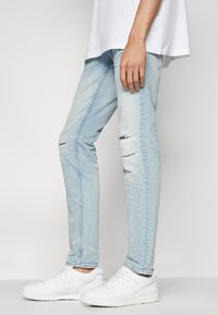 Jeans ajustados de color azul claro con parches rasgados en las rodillas y una textura suave, combinados con zapatillas deportivas blancas. Diseño casual y moderno.