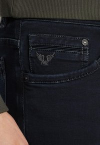 Jean en denim bleu foncé avec un petit logo d'aigle brodé sur la poche arrière, des coutures détaillées et un bouton en métal.