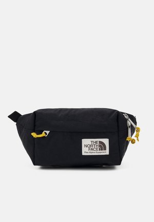 Sac banane rectangulaire noir avec des tirettes de fermeture éclair jaunes et un patch logo blanc "The North Face Fine Alpine Equipment" à l'avant.