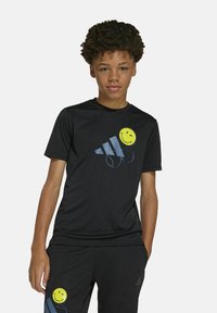 Camiseta deportiva negra con una cara sonriente amarilla y rayas diagonales azules. Mangas cortas, material ligero, textura suave.
