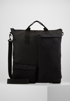 Bolso tote rectangular negro con dos bolsillos frontales con cremallera, asas superiores y una correa de hombro ajustable, colocado sobre una superficie blanca.