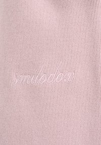 Tissu rose clair côtelé avec du texte cursif brodé légèrement en relief, lisant "smilodox" en fil assorti.