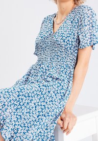 Robe fleurie bleue à manches courtes, col en V et corsage froncé. Présente des motifs de fleurs blanches sur toute la surface. Tissu doux et léger.