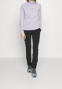 Parte superior de cremallera de manga larga en color púrpura claro con diseño texturizado; pantalones ajustados negros con bolsillo lateral con cremallera; zapatos deportivos grises y naranjas.