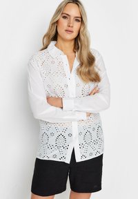 Camisa blanca de botones con patrón de ojales florales, mangas largas y cuello clásico. Combinada con pantalones cortos negros. Textura de tela suave.