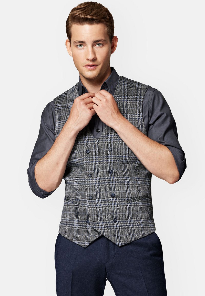 Szary kamizelka w kratę z podwójnym zapięciem, z niebieskimi akcentami i czarnymi guzikami, noszona na ciemnej koszuli. Gładka tekstura materiału.