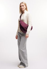 Borsa a tracolla color bordeaux realizzata in tessuto morbido, caratterizzata da un design curvo e una tracolla regolabile. Abbinata a un maglione leggero e a jeans a gamba larga grigi.