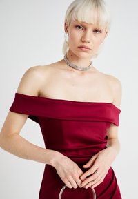 Robe bordeaux à épaules nues avec une silhouette ajustée, une texture lisse et un détail drapé. Accentuée par un collier en argent et tenant un sac à main circulaire.