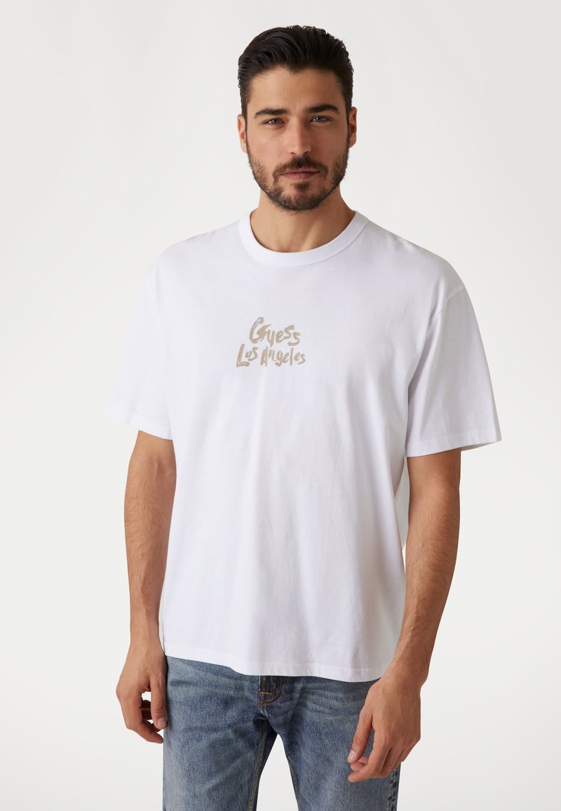 Hvid bomulds t-shirt med et struktureret beige logo med teksten "Guess Los Angeles" på forsiden, med en klassisk rund hals og afslappet pasform.