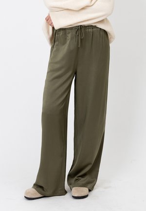 Personne portant un pantalon ample vert olive avec cordon de serrage et des chaussons beiges, avec un pull crème partiellement visible.