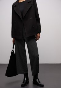 Personne portant un manteau noir croisé, un pantalon large et foncé, des bottes noires, tenant un sac à main noir sur un fond uni.