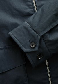 Dark blue jacket detail featuring a buttoned cuff, zip closure, and textured fabric with a slightly glossy finish.