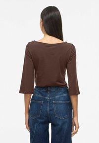 Sieviete ar gariem taisniem matiem, valkājot brūnu topu ar trīs ceturtdaļu piedurknēm un zilas džinsa bikses ar augstu jostasvietu, redzama no muguras.