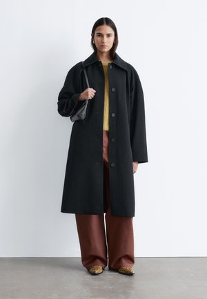 Frau steht vor weißer Wand, trägt einen langen schwarzen Mantel mit Knöpfen, gelben Pullover, weite braune Hose und hält eine schwarze Umhängetasche.