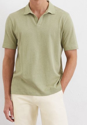 Mann trägt hell olivgrünes kurzärmeliges Poloshirt und off-white Hose, steht vor weißem Hintergrund.