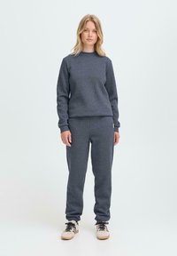 Kvinne som står iført en mørkegrå hettegenser og matchende joggebukser med beige og svarte joggesko mot en lys bakgrunn.