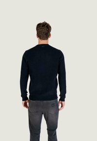 Tamno plavi pulover s rebrastom teksturom, dugim rukavima i okruglim izrezom, uparen s sivim farmerkama koje imaju detalj na stražnjem džepu.