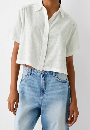 Femme portant une chemise courte blanche à manches courtes et un jean taille haute bleu clair sur un fond blanc uni.