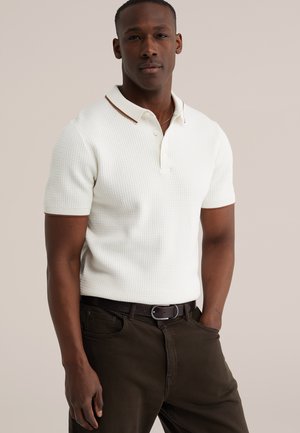 Mężczyzna w białej, teksturowanej koszulce polo z krótkim rękawem i brązową lamówką, ciemnym paskiem i ciemnych spodniach, stojący z jedną ręką w kieszeni.