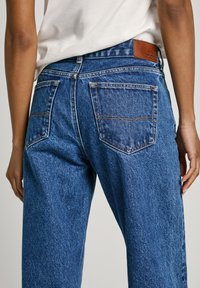 Kõrge vöökohaga sinised denim püksid, millel on klassikaline lõike, kaks tagakotti ja pruun nahklogopatch vöökohal.