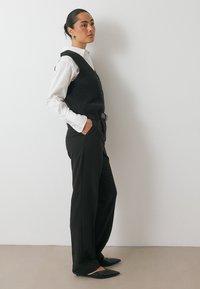 Chaleco de canalé negro sobre una camisa blanca de botones, combinado con pantalones de pierna ancha negros y zapatos planos de punta. Diseño minimalista, esquema de color monocromático.