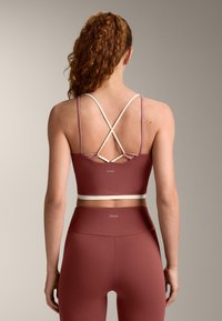 Donna con capelli lunghi e ricci che indossa un reggiseno sportivo color vinaccia con spalline beige incrociate e leggings a vita alta abbinati, vista da dietro.