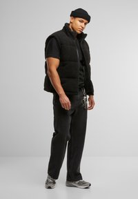 Black fleece vest with a high collar, worn over a black t-shirt. Dark jeans, silver keys on a chain, and grey athletic shoes complete the look.