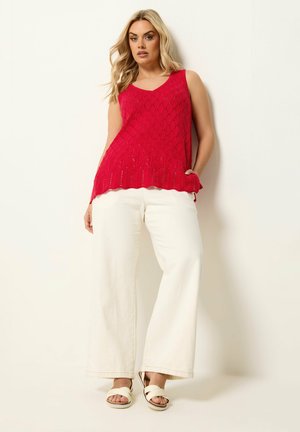 Blonde Frau trägt ein rotes ärmelloses Stricktop, weiße weit geschnittene Hose und weiße Sandalen, steht vor einem schlichten hellen Hintergrund.