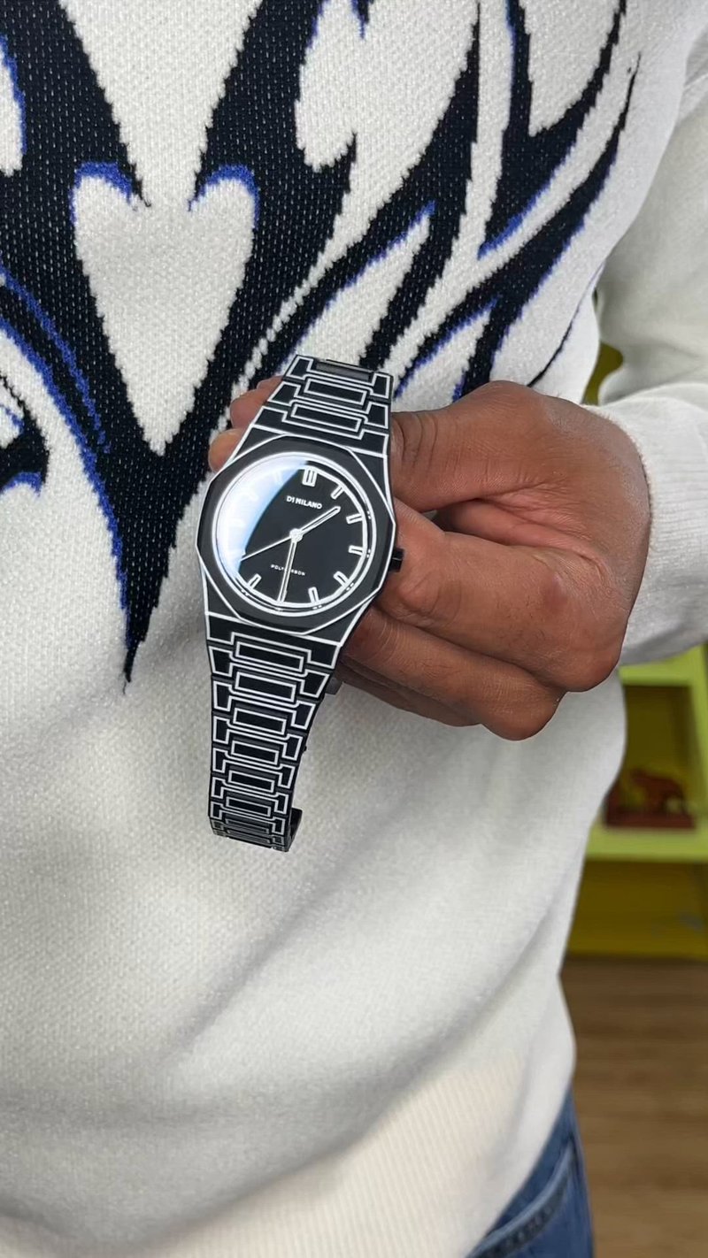 Main tenant une montre-bracelet à motif noir et blanc avec un cadran noir et des marqueurs argentés, sur un pull blanc avec un motif noir et bleu.