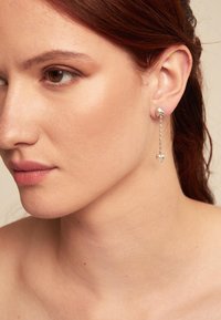 Pendientes de plata con un diseño de aro y un colgante en forma de corazón suspendido por una cadena, que muestra metal pulido y detalles elegantes.