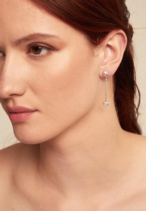 Pendientes de plata con un diseño de aro y un colgante en forma de corazón suspendido por una cadena, que muestra metal pulido y detalles elegantes.