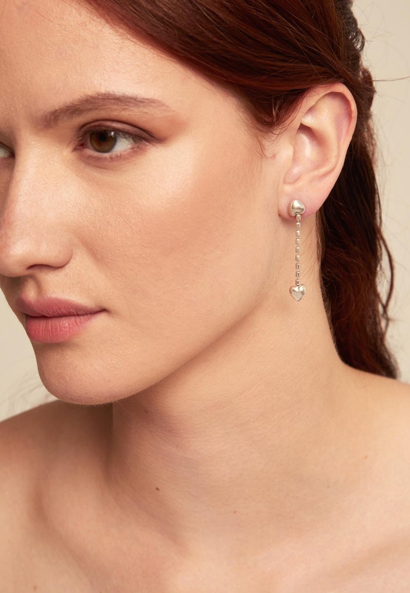 Pendientes de plata con un diseño de aro y un colgante en forma de corazón suspendido por una cadena, que muestra metal pulido y detalles elegantes.