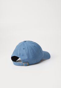 Casquette de baseball en coton bleu avec une sangle ajustable et une boucle en laiton posée sur une surface blanche, vue depuis le côté arrière gauche.