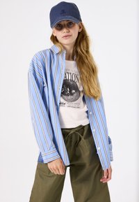 Helles blau gestreiftes Hemd mit Knöpfen, weißes T-Shirt mit Grafik und lockere grüne Hose. Abgerundet mit einer marineblauen Kappe. Lässiges und einfaches Design.