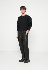 Čierna mikina s malým červeným logom, kombinovaná s čiernymi koženými cargo nohavicami s viditeľnými vreckami a hladkou textúrou. Čierne tenisky s pruhmi.