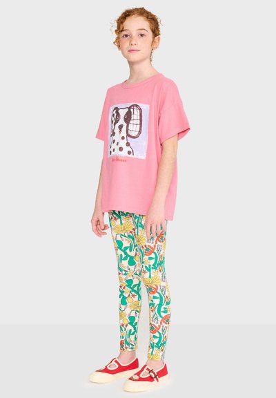 Enfant aux cheveux roux bouclés portant un t-shirt ample rose avec un motif de chien, des leggings à motif floral et des chaussures rouges à enfiler avec des semelles blanches.