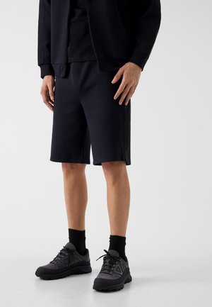 Persona in piedi con pantaloncini neri, scarpe sportive nere con calze nere e una giacca nera con zip, davanti a uno sfondo chiaro e semplice.