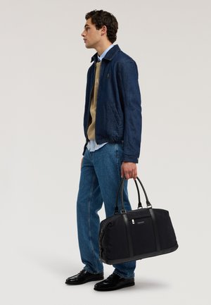 Młody mężczyzna w niebieskiej kurtce i dżinsach trzymający dużą czarną torbę, stojący bokiem na jednolitym jasnym tle.