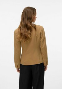 Blazer marron sur mesure avec des manches longues, coupe structurée et coutures arrière. S'associe à un pantalon noir, le tout sur un fond clair.