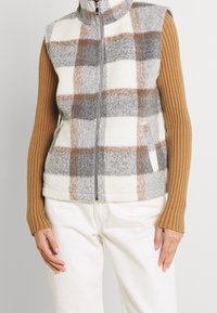 Fleece-Weste mit hohem Kragen im Karomuster in Grau, Braun und Creme, vorne geöffnet, getragen über einem gerippten Pullover.