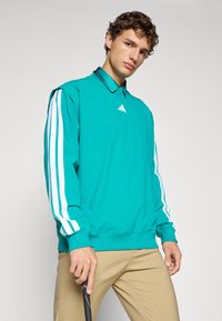 Young man wearing turquoise Adidas sweatshirt with white stripes on sleeves and holding a black golf club against a gray background.
