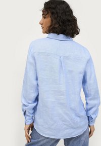 Camisa de manga larga azul claro hecha de una tela texturizada, con un canesú en la parte posterior, costuras en los hombros y puños abotonados. Dobladillo redondeado.