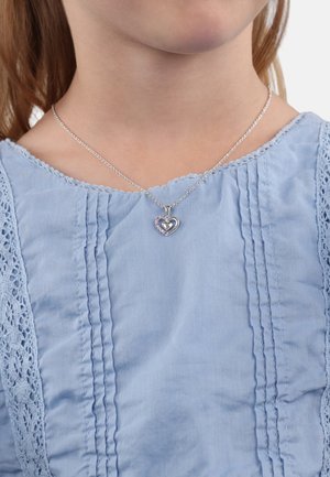 Silbernes Herz-Halsband mit einem kleineren Herz, das mit rosa Steinen eingelegt ist, getragen über einer hellblauen Bluse mit Spitze und vertikalen Falten.