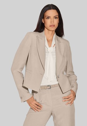 Beige, taillierter Blazer mit verkürzter Passform, strukturierten Schultern und einem einzigen Knopfverschluss, kombiniert mit einer weißen Bluse und beigen Hosen.