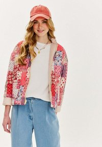 Farebná patchworková bunda s kvetinovými a pruhovanými vzormi, spojená s svetlým topom, džínsovými nohavicami a ružovou čiapkou.